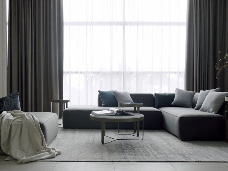 prikaz sive ugaone sa jastucima, sive fotelje i ćebeta preko nje, tepiha na sredini sobe i stolčića