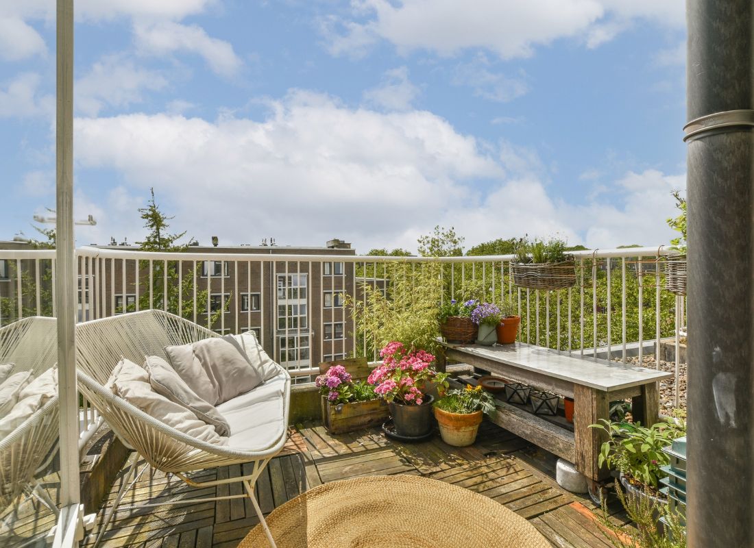 Udobna terasa ima šarene saksije sa cvećem i udobnu stolicu, okružena zelenilom i plavim nebom, savršena za opuštanje i uživanje u prirodi.